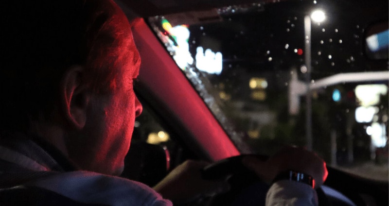 Older man drives his car at night, a red stoplight reflects on his face.